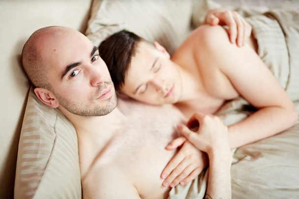 Joven hombre calvo mirando a la cámara mientras está acostado en la cama y abrazando a su pareja gay durmiendo
 - Foto, imagen