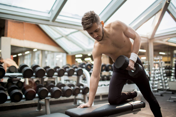Joven hombre sin camisa apoyado en el brazo derecho y la rodilla doblada mientras hace ejercicio con pesadas mancuernas en el gimnasio
 - Foto, imagen