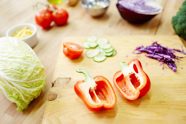 Дві половини свіжого червоного паприки на дерев'яній дошці серед інших інгредієнтів для овочевого салату або рагу
 - Фото, зображення