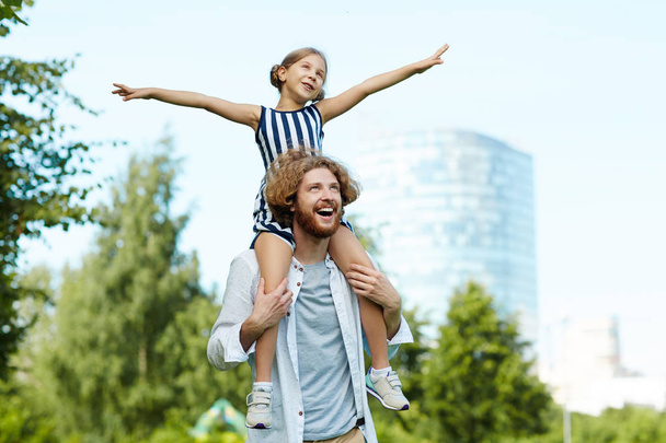 Menina feliz com braços estendidos sentados nos ombros do pai durante a caminhada no parque
 - Foto, Imagem