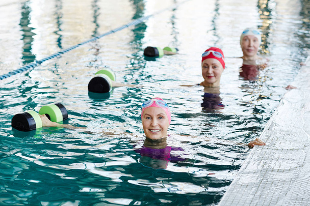 Riga di donne anziane felici in costume da bagno che allungano le braccia destre mentre sollevano i bilancieri durante l'allenamento in acqua
 - Foto, immagini