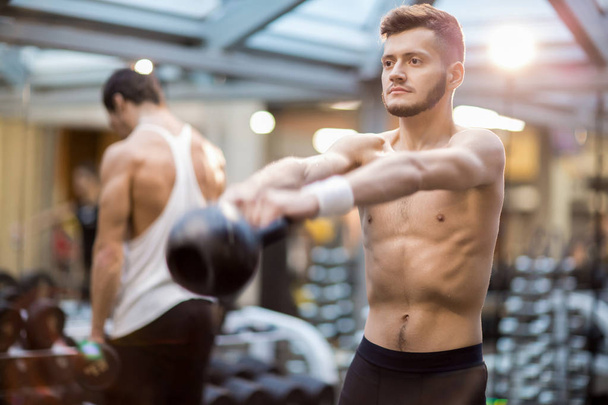 Без сорочки чоловік з розтягнутими руками піднімає важкий чайник під час тренувань у сучасному спортивному клубі
 - Фото, зображення