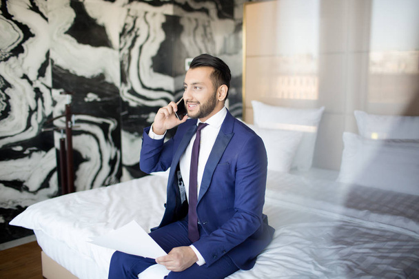 Jonge vrolijke econoom in formalwear zittend op bed in de hotelkamer en in gesprek met klanten op smartphone - Foto, afbeelding