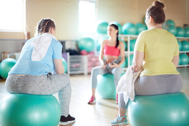 Dos mujeres jóvenes sobre tamaño en ropa deportiva sentadas en fitballs delante de su instructor de fitness
 - Foto, imagen