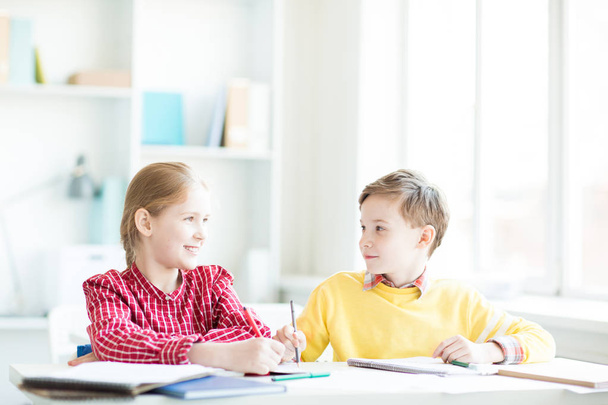 Dois pequenos alunos amigáveis sentados à mesa em sala de aula, pensando em ideias e discutindo-as
 - Foto, Imagem