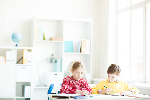 Двоє маленьких однокласників сидять за столом, малюють або роблять нотатки в своїх книгах на уроці
 - Фото, зображення