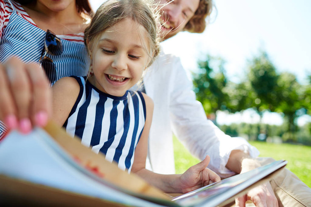 Onnellinen pieni tyttö ja hänen vanhempansa lukevat kirjaa viettäessään aikaa puistossa kesäviikonloppuna
 - Valokuva, kuva