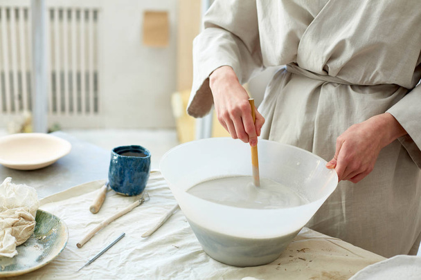 Jeune femme artisanale créative mélange de poudre d'argile avec de l'eau dans un bol en plastique sur la table
 - Photo, image