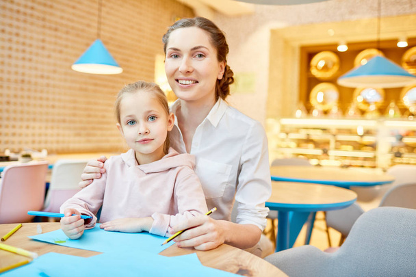 Молодая женщина и маленькая девочка с карандашами смотрят в камеру, сидя за столом в кафе на досуге
 - Фото, изображение