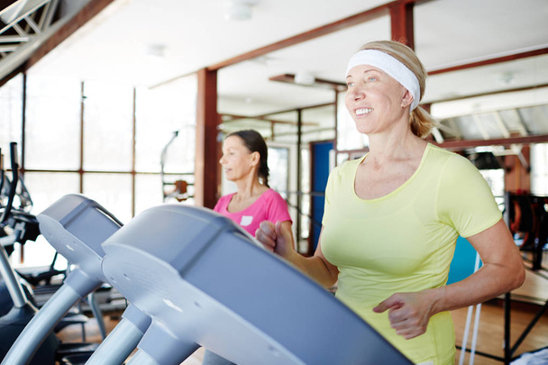 Happy aged woman running on treadmill in sports center with her friend on background - Photo, Image