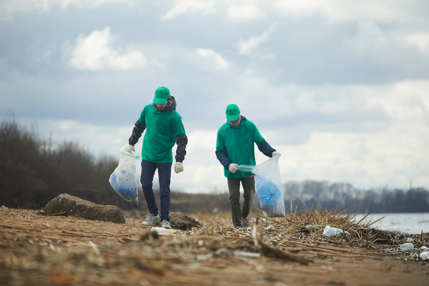 Two men with big sacks walking along river bank, picking up litter and putting it in those sacks - Фото, зображення