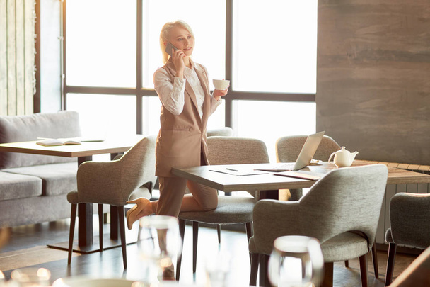 Young elegant businesswoman with cup of tea or coffee standing by table in cafe, talking by smartphone and making new appointment with one of clients - Foto, Bild