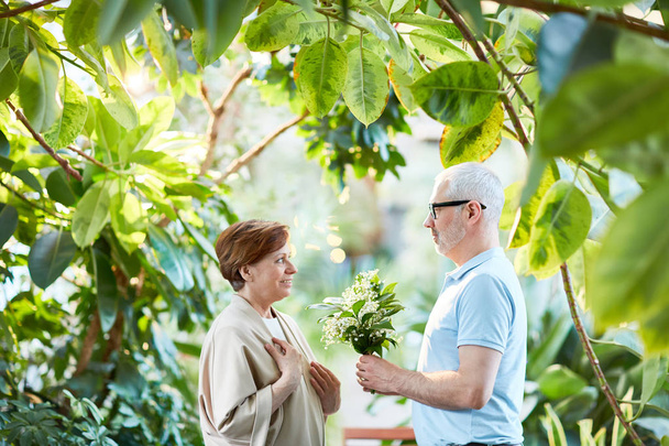 Hombre maduro dando ramo de lirios frescos y aromáticos del valle a su esposa mientras pasea por el jardín
 - Foto, Imagen