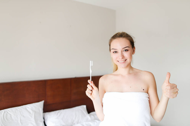 Mladá milá žena zabalená do bílého ručníku, stojící s zubní kartáčkem blízko postele, ukazující palce. - Fotografie, Obrázek