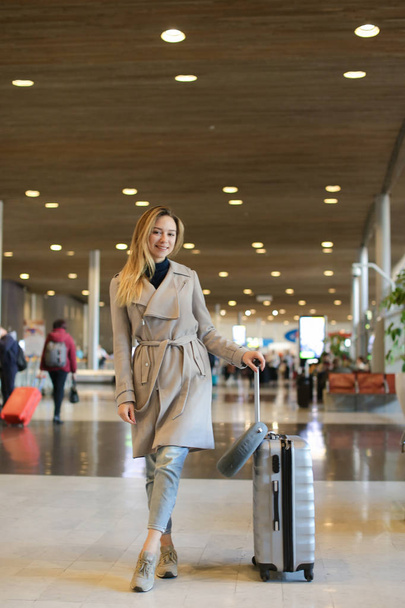 Γυναικείο πρόσωπο στέκεται στην αίθουσα αναμονής αεροδρομίου, με βαλίτσα και φορώντας γκρι παλτό. - Φωτογραφία, εικόνα