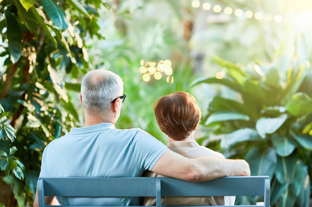Vista trasera de la pareja mayor cariñosa sentada en el banco en el jardín o el parque en el ocio
 - Foto, Imagen
