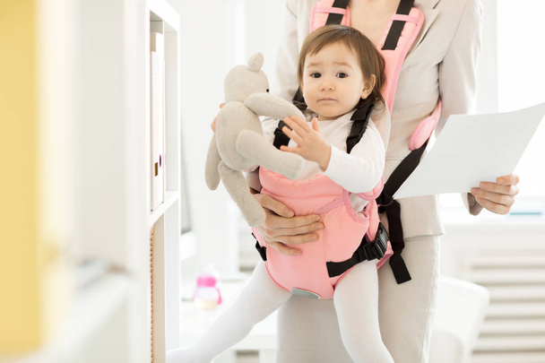 Baby girl with teddybear sitting in pink carrier in front of her busy mother working with papers in office - Photo, Image