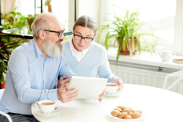 Contemporary senior couple with tablet and earphones talking through video-chat while sitting in cafe by cup of tea with cookies - Photo, Image