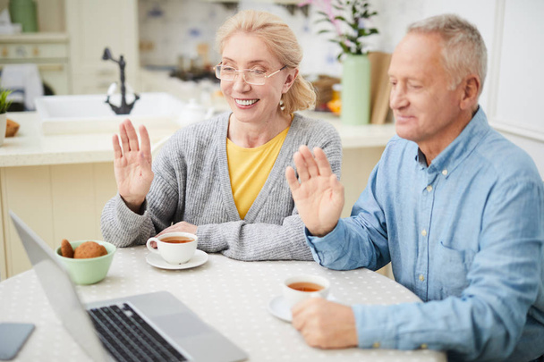 Szczęśliwy dziadków, siedzi przy stole w kuchni i machanie rękami do swoich znajomych lub krewnych, podczas rozmowy przez czat wideo - Zdjęcie, obraz