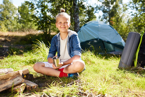 Happy boy with crossed legs sitting on grass and having tea by campfire on sunny morning - Photo, Image