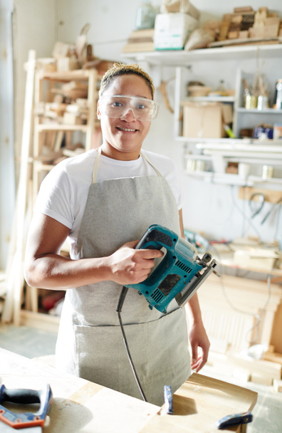 Female carpenter with electric fretsaw looking at camera through protective eyeglasses in workroom - Photo, Image