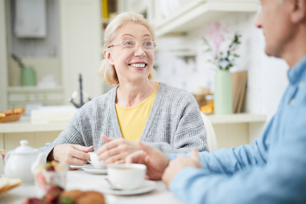 Smiling mature woman having talk to her husband by cup of tea in the kitchen - Foto, immagini