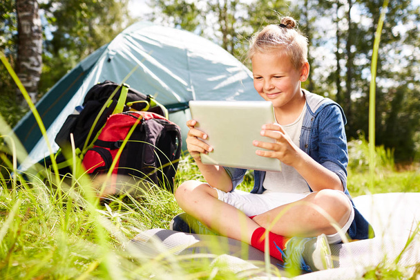 Kleiner Backpacker mit Touchpad sitzt auf grünem Gras und schaut am Nachmittag einen Online-Film mit seinem Zelt im Hintergrund - Foto, Bild