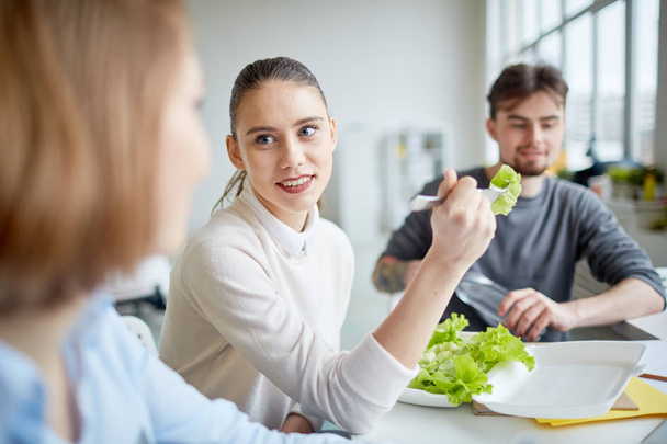 Молодая улыбающаяся деловая женщина ест салат за столом со своими коллегами в офисе
 - Фото, изображение