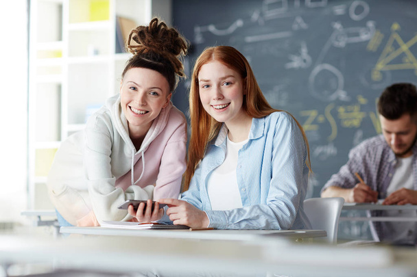 zwei glückliche Mädchen in Freizeitkleidung, die lächelnd in die Kamera schauen, während sie sich zwischen den Unterrichtsstunden im College unterhalten - Foto, Bild