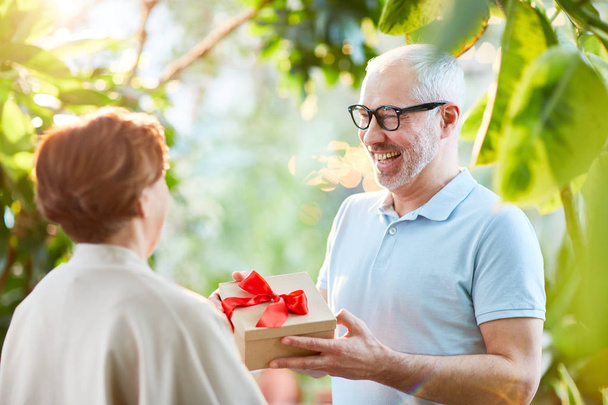 Ευτυχής ανώτερος άνθρωπος, δίνοντας τη σύζυγό του κουτί με δώρο για την επέτειο του γάμου τους κατά τη διάρκεια του περιπάτου στον κήπο - Φωτογραφία, εικόνα