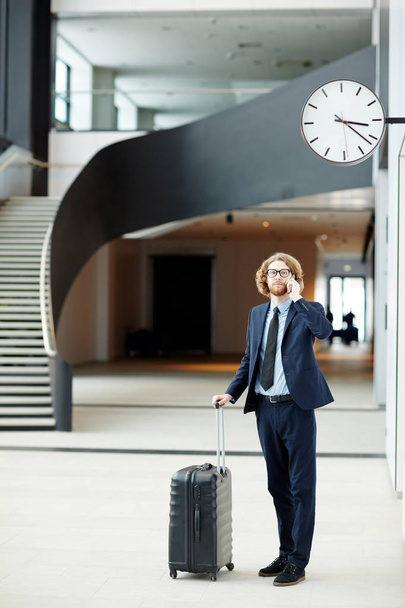 Joven viajero de negocios con equipaje hablando por teléfono inteligente mientras espera la salida
 - Foto, Imagen