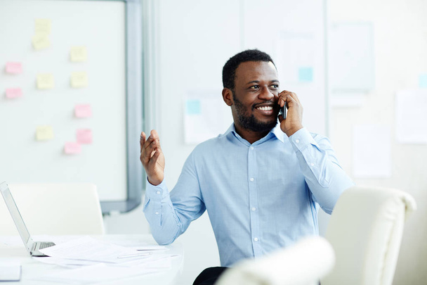 Afrikanisch-amerikanischer Geschäftsmann in Formalbekleidung spricht während seiner Arbeit im Büro mit dem Kunden per Smartphone - Foto, Bild