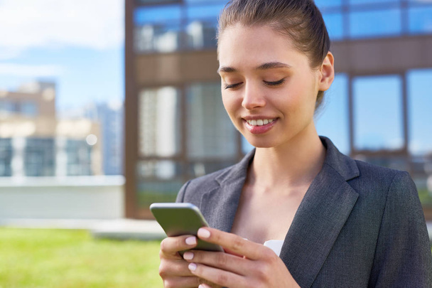 Gelukkig zakenvrouw in formalwear communiceren via een mobiele boodschappers in haar smartphone in stedelijke omgeving - Foto, afbeelding