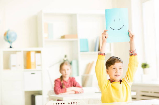 carino scolaro alzando la mano tenendo blu carta con disegnato sorriso mentre rispondendo a insegnante
 - Foto, immagini