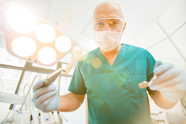 Зрелый стоматолог в форме и защитной маске и очках держит дрель и зеркало
 - Фото, изображение