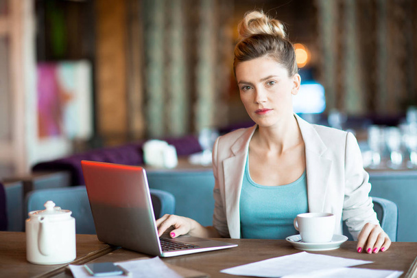 Молодая уверенная деловая женщина смотрит в камеру, сидя за столом и ища в сети
 - Фото, изображение