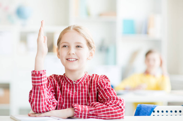 Écolière avec toothy sourire levant la main tout en étant assise à son bureau à la leçon
 - Photo, image