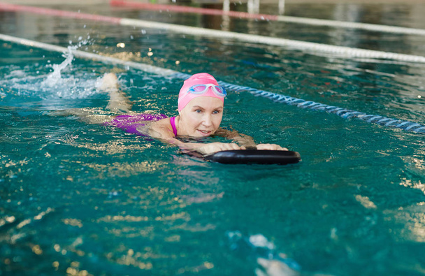 Donna anziana in costume da bagno tenuta da anello di gomma nera mentre nuota in avanti in piscina
 - Foto, immagini