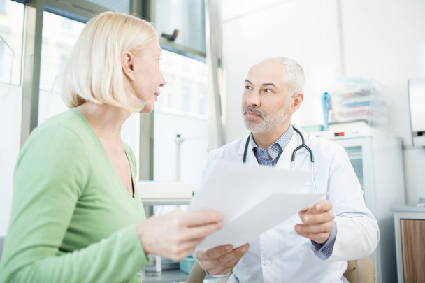 Confident clinician and his patient discussing medical papers or prescriptions during appointment - Foto, afbeelding