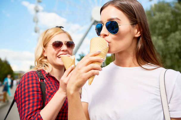Two stylish girls in sunglasses eating tasty icecream in amusement park on background of blue sky and ferris wheel - Fotó, kép