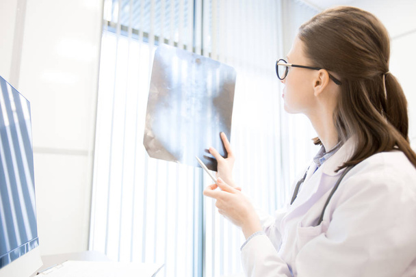 Jovem radiologista em uniforme olhando para a imagem de raios-x enquanto trabalhava em hospital ou clínicas
 - Foto, Imagem