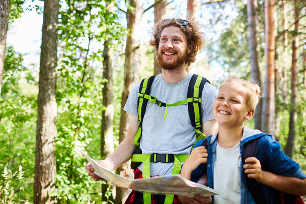 Χαρούμενα backpackers με χάρτη που ψάχνετε μέρος για να εγκατασταθούν ενώ το περπάτημα μέσα από δάσος σε καλοκαιρινή μέρα - Φωτογραφία, εικόνα