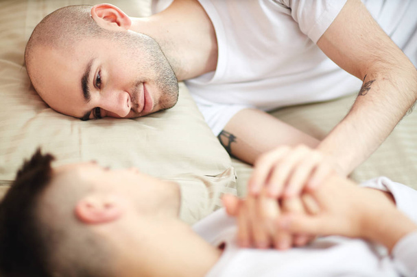 ベッドでリラックスしながら彼のパートナーを見て若いロマンチックなゲイの男性 - 写真・画像