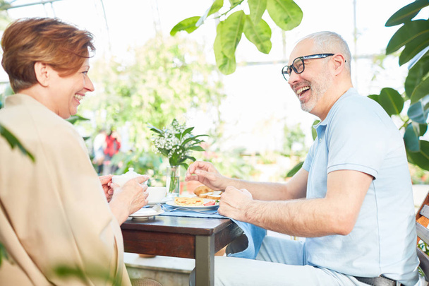 lachende Seniorin und Ehemann sitzen beim Frühstück am Tisch und unterhalten sich - Foto, Bild