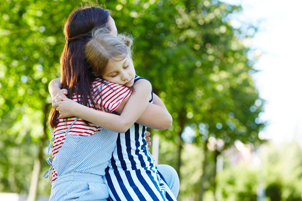 Menina feliz sentado em seu joelho mãe ao abraçá-la no parque no dia de verão
 - Foto, Imagem