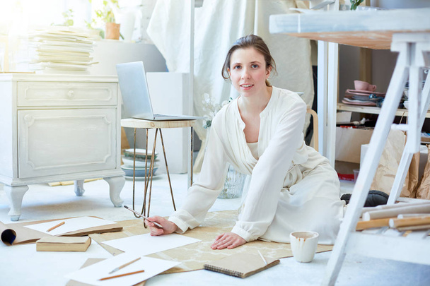 Chica creativa en vestido blanco bosquejando en papeles mientras trabaja en el suelo de su estudio
 - Foto, Imagen