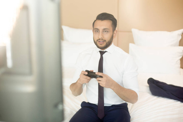 Επιχειρηματίας στην formalwear, κάθεται στο κρεβάτι μπροστά από την οθόνη και το τυχερό παιχνίδι σε δωμάτιο ξενοδοχείου στο διάλειμμα - Φωτογραφία, εικόνα
