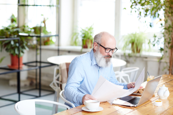 Rušné starší muž hledá prostřednictvím finanční doklady při sezení u stolu v kavárně s notebookem v popředí - Fotografie, Obrázek