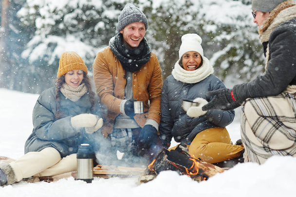 Радісна молодь в зимовому одязі, сидячи в снігу, збуджує вогонь і гарячі напої
 - Фото, зображення