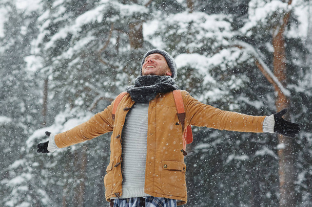 Happy young man with backpack enjoying snowfall while outstretching his arms and closed eyes - Photo, Image